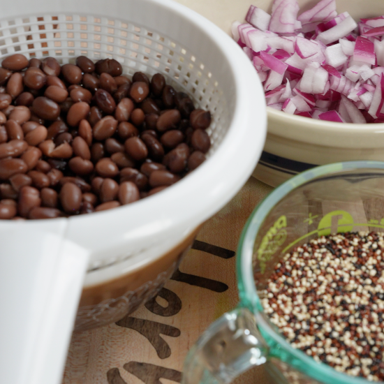 Drained black beans in a colander, chopped onion and garlic, and quinoa in a measuring cup.